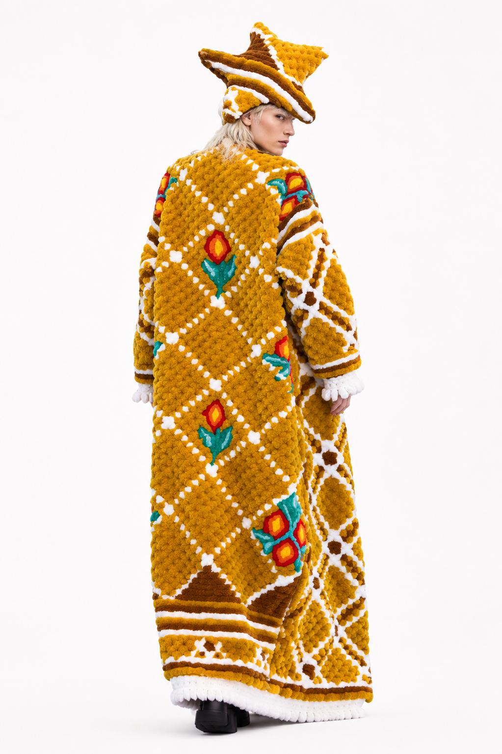 A model is shown from the back wearing a long, mustard yellow knit coat with a white diamond lattice pattern, colorful floral motifs, and a matching pointed yellow hat against a plain white background.